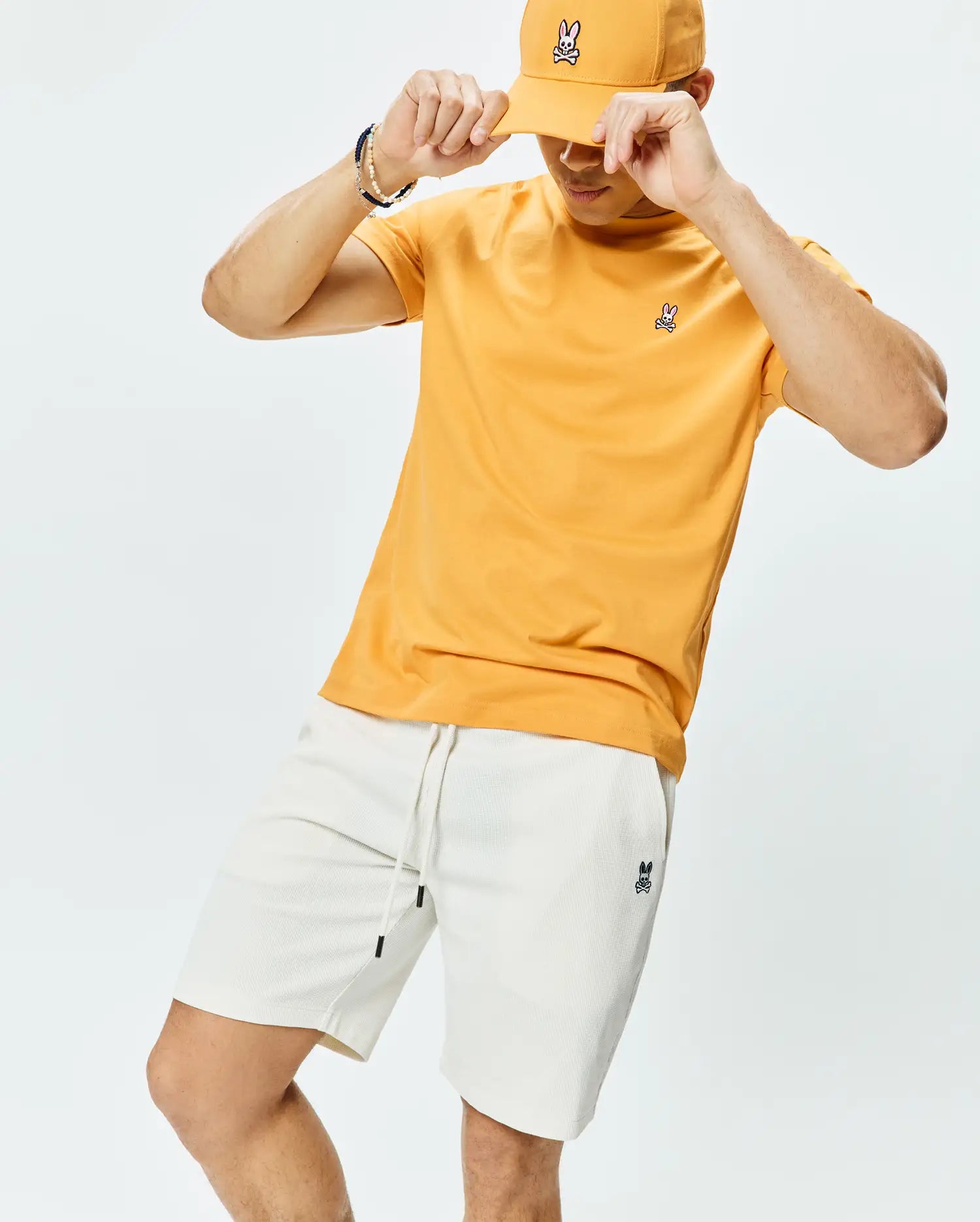 Orange Mens Classic Crew Neck Tee with a small embroidered Psycho Bunny skull logo on the left chest, cream textured shorts with a matching logo, and an orange Psycho Bunny baseball cap.