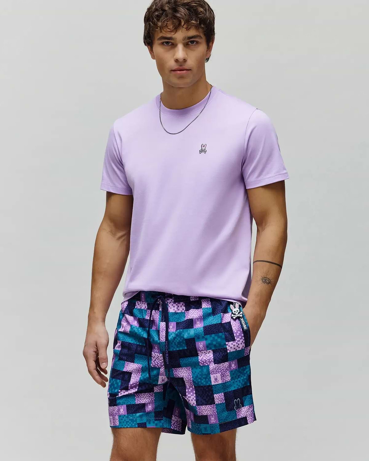 Mens Classic Crew Neck Tee, a classic fit light purple jersey t-shirt with small embroidered Psycho Bunny logo, paired with purple and teal geometric patchwork shorts featuring multiple bunny skull logos.