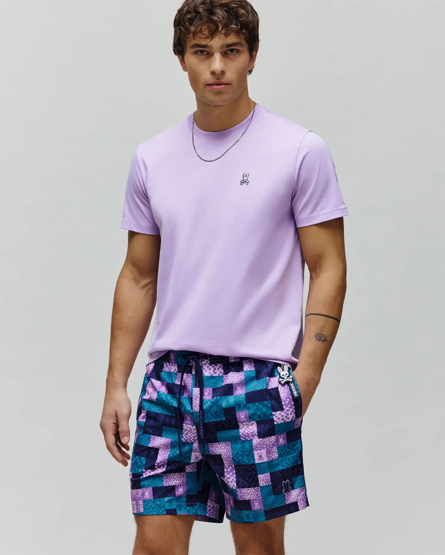 Mens Classic Crew Neck Tee, a classic fit light purple jersey t-shirt with small embroidered Psycho Bunny logo, paired with purple and teal geometric patchwork shorts featuring multiple bunny skull logos.