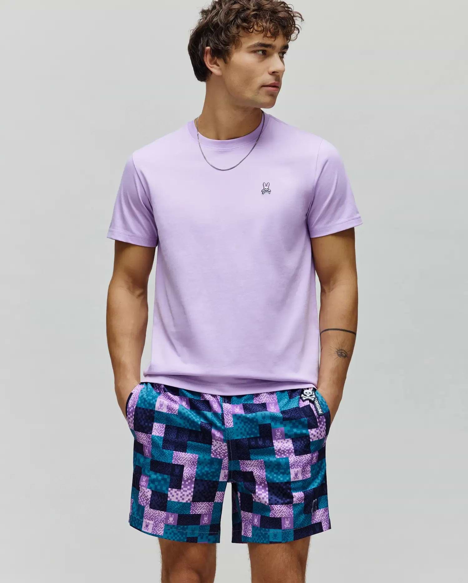 Mens Classic Crew Neck Tee, lavender jersey, with embroidered Psycho Bunny skull logo on left chest. Paired with relaxed shorts in a teal and purple pixelated patchwork pattern with repeated Psycho Bunny logos.