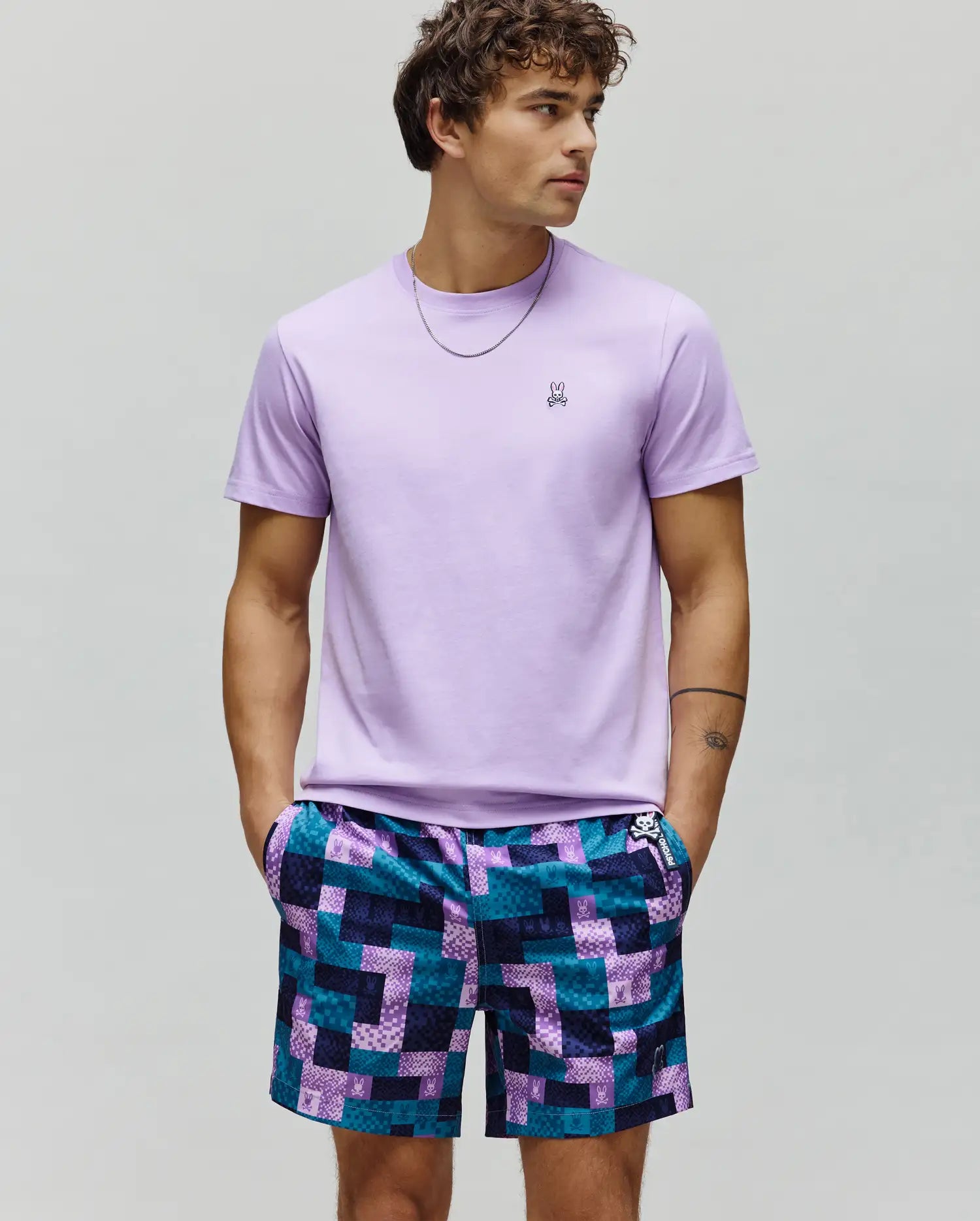 Mens Classic Crew Neck Tee, lavender jersey, with embroidered Psycho Bunny skull logo on left chest. Paired with relaxed shorts in a teal and purple pixelated patchwork pattern with repeated Psycho Bunny logos.
