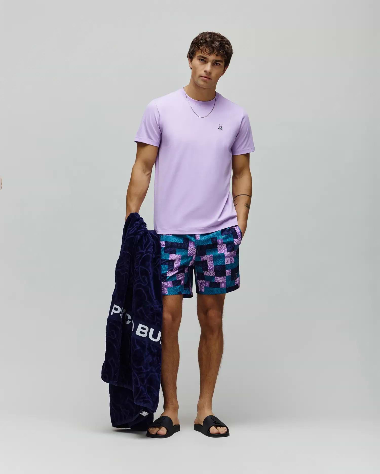 Mens Classic Crew Neck Tee, a classic fit lavender jersey t-shirt with an embroidered chest Psycho Bunny logo, paired with relaxed geometric print swim shorts in navy, teal, and purple.