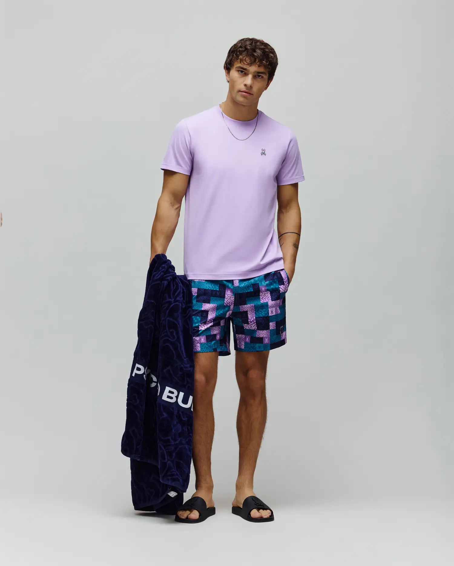 Mens Classic Crew Neck Tee, a classic fit lavender jersey t-shirt with an embroidered chest Psycho Bunny logo, paired with relaxed geometric print swim shorts in navy, teal, and purple.