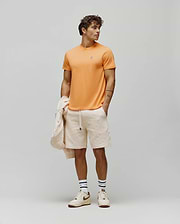 Mens Classic Crew Neck Tee in vibrant orange jersey with a small embroidered Psycho Bunny skull logo on the left chest, paired with cream relaxed fit shorts featuring an allover embossed bunny skull pattern.