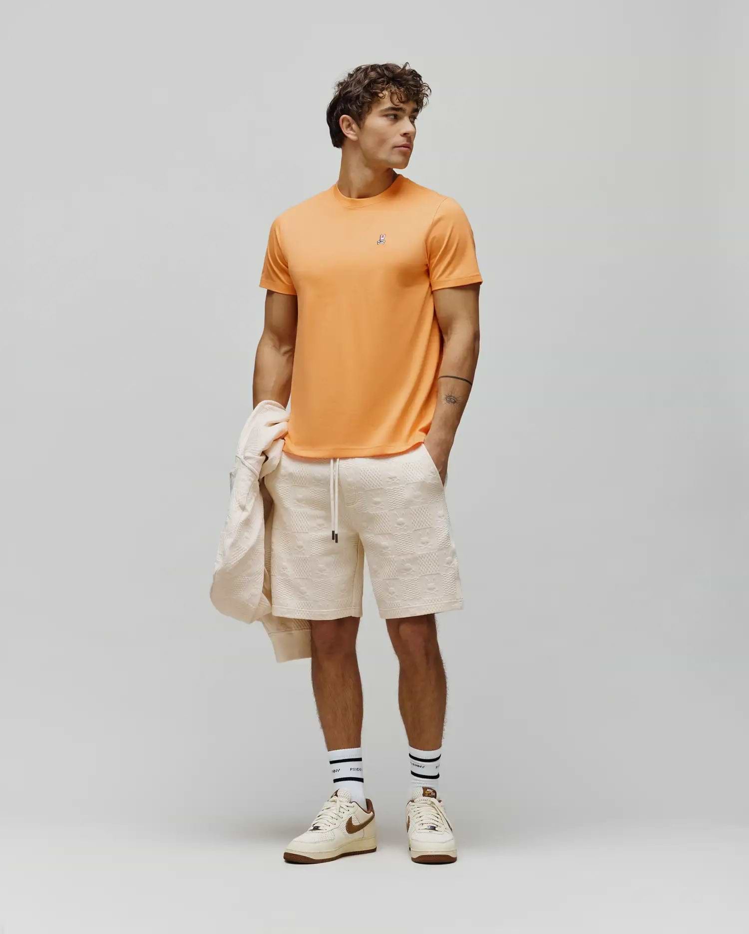 Mens Classic Crew Neck Tee in vibrant orange jersey with a small embroidered Psycho Bunny skull logo on the left chest, paired with cream relaxed fit shorts featuring an allover embossed bunny skull pattern.