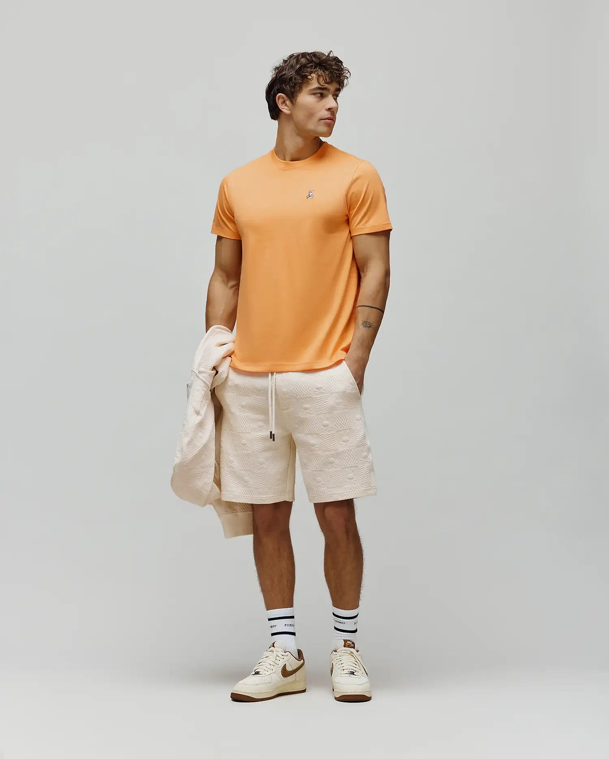 Mens Classic Crew Neck Tee in vibrant orange jersey with a small embroidered Psycho Bunny skull logo on the left chest, paired with cream relaxed fit shorts featuring an allover embossed bunny skull pattern.
