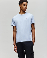 A young man stands against a gray background, wearing Psycho Bunny's MENS CLASSIC CREW NECK TEE (B6U014F200) in light blue Pima cotton with a small chest logo, paired with black cargo pants.