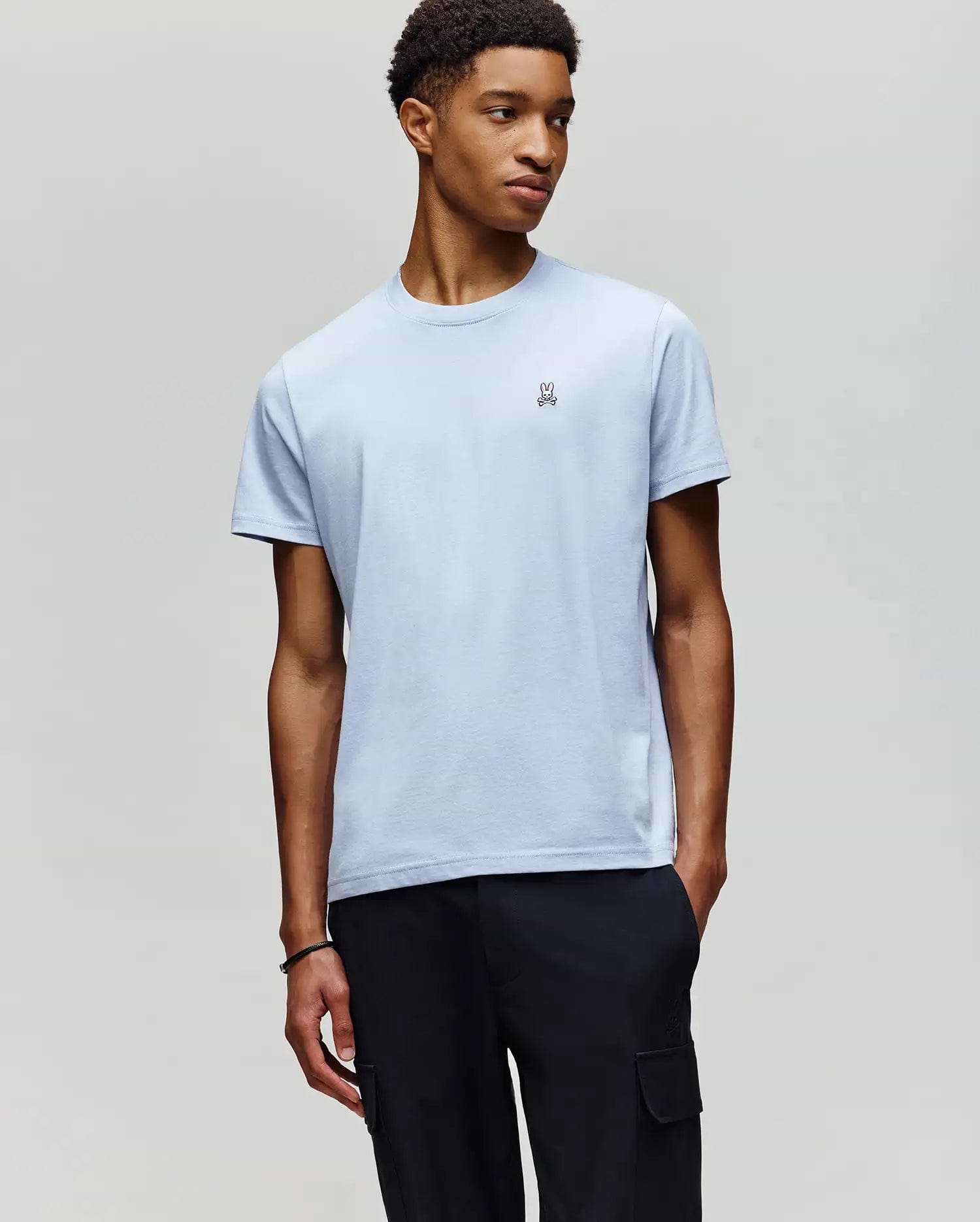 A young man stands against a gray background, wearing Psycho Bunny's MENS CLASSIC CREW NECK TEE (B6U014F200) in light blue Pima cotton with a small chest logo, paired with black cargo pants.