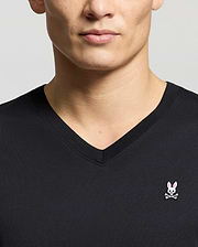 A man wears a Psycho Bunny MENS CLASSIC V NECK TEE (B6U100ARPC), made from premium Pima cotton, featuring a pink bunny head and white eyes above bold crossbones on a light gray background.