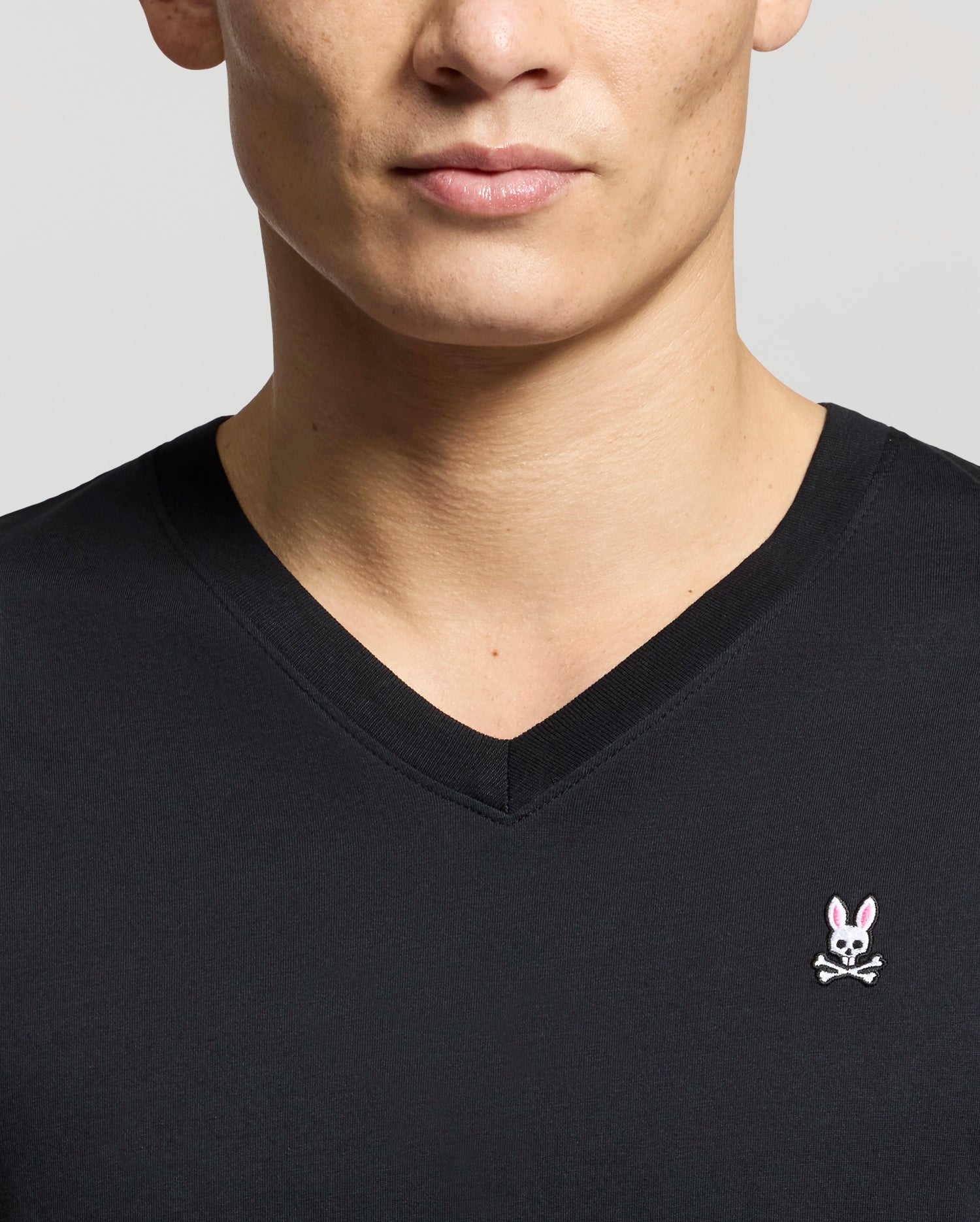 A man wears a Psycho Bunny MENS CLASSIC V NECK TEE (B6U100ARPC), made from premium Pima cotton, featuring a pink bunny head and white eyes above bold crossbones on a light gray background.