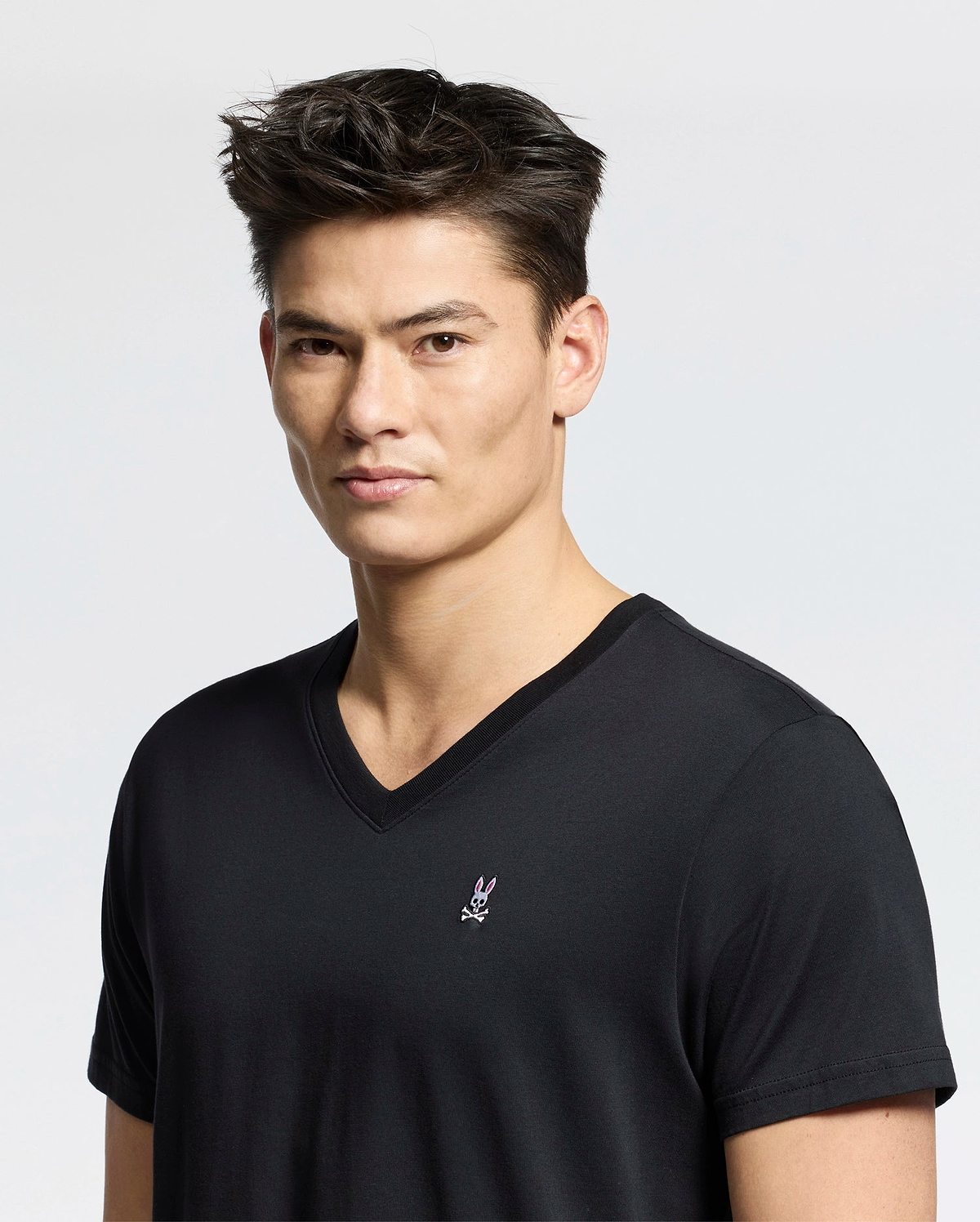 Someone wears a Psycho Bunny MENS CLASSIC V NECK TEE - B6U100ARPC in black, made from soft Pima cotton, with a small logo featuring a rabbit with crossed bones on the chest. The background is plain gray.