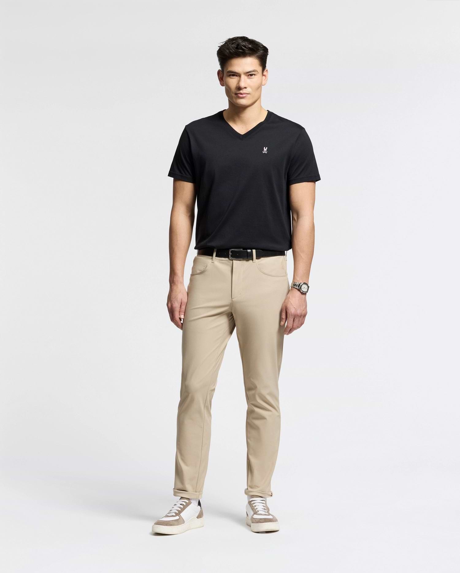 Against a white backdrop, a man models effortless style wearing Psycho Bunny's MENS CLASSIC V NECK TEE (B6U100ARPC) in black. He pairs it with beige pants, white sneakers with black details, and a wristwatch. With short dark hair and relaxed posture, he exudes laid-back elegance.