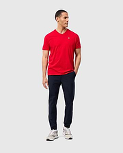 A person wearing a red Psycho Bunny MENS CLASSIC V NECK TEE (B6U100CRPC), paired with navy pants and white sneakers, stands against a plain background. They have short dark hair, are gazing to the side, and have one hand in their pocket.