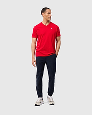 A person wearing a red Psycho Bunny MENS CLASSIC V NECK TEE (B6U100CRPC), paired with navy pants and white sneakers, stands against a plain background. They have short dark hair, are gazing to the side, and have one hand in their pocket.