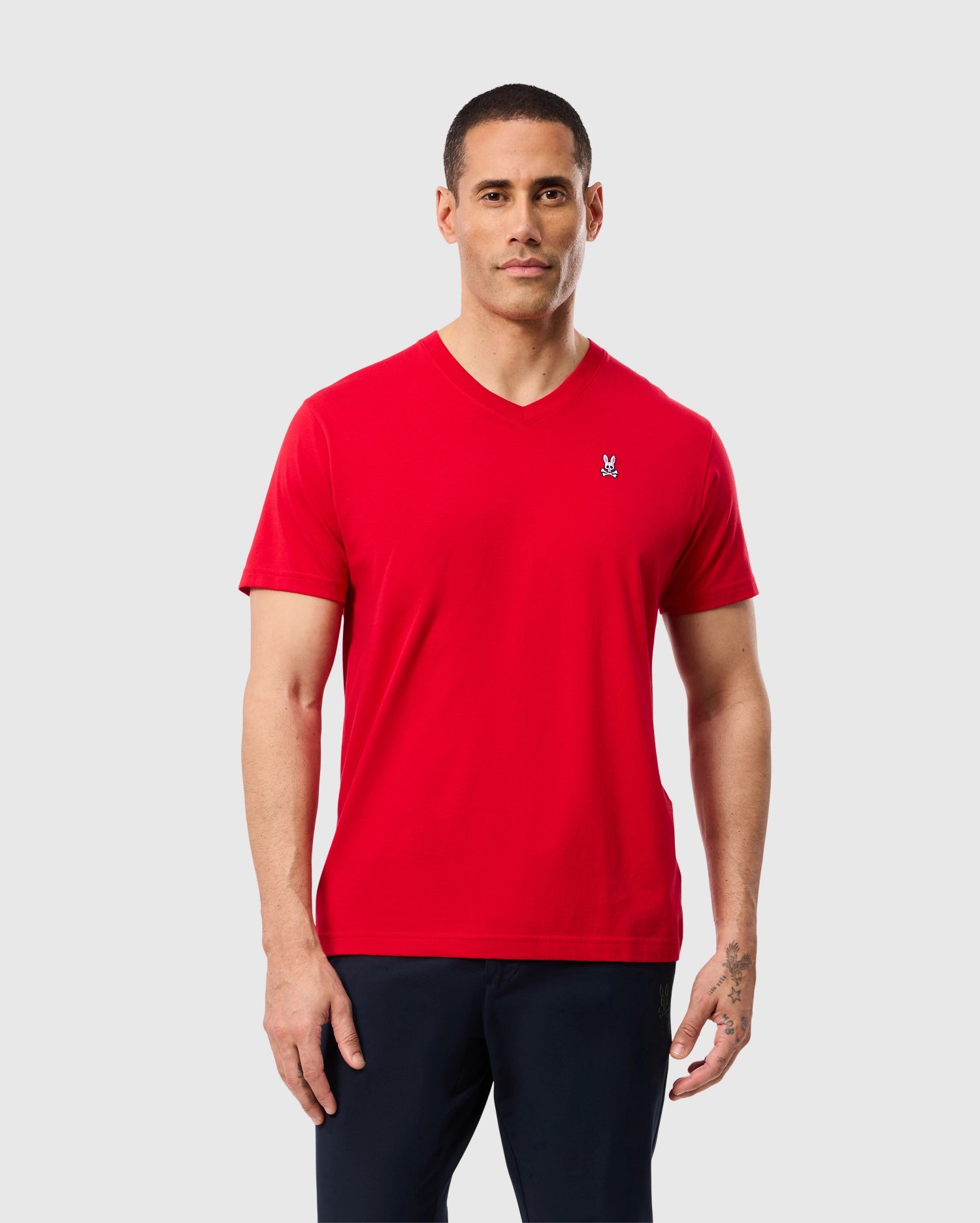 A person wearing the MENS CLASSIC V NECK TEE - B6U100CRPC by Psycho Bunny, which features a subtle logo embroidered on the chest and is crafted from soft Pima cotton, stands against a plain light gray background in black pants.