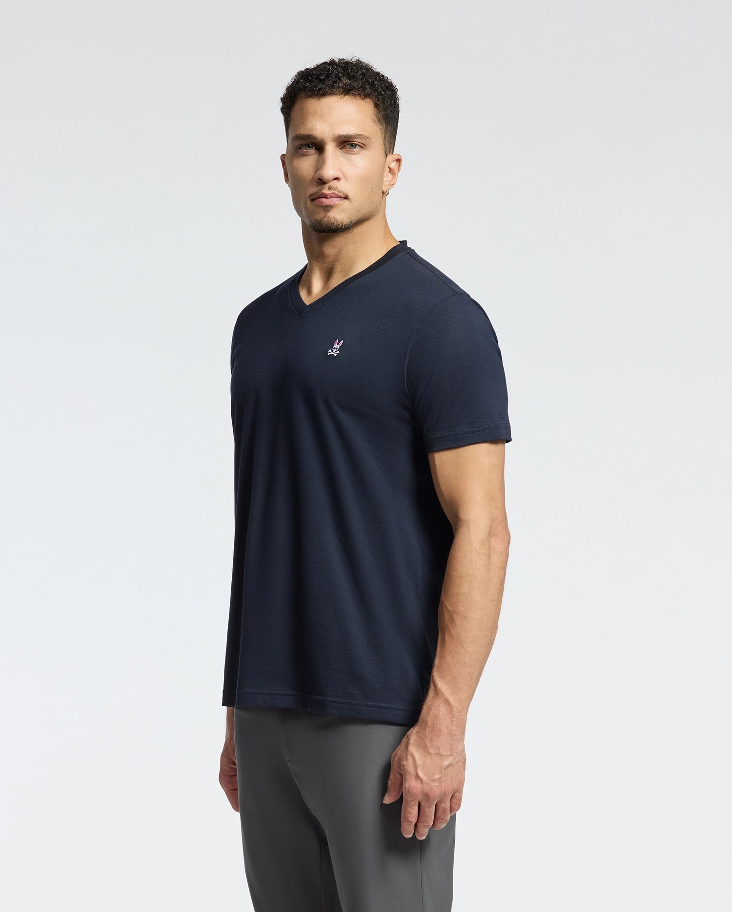 A man with short curly hair is wearing a dark Psycho Bunny MENS CLASSIC V NECK TEE - B6U100ARPC and gray pants. He stands against a plain white background, gazing confidently at the camera. A small logo is visible on the left side of the Pima cotton T-shirt.