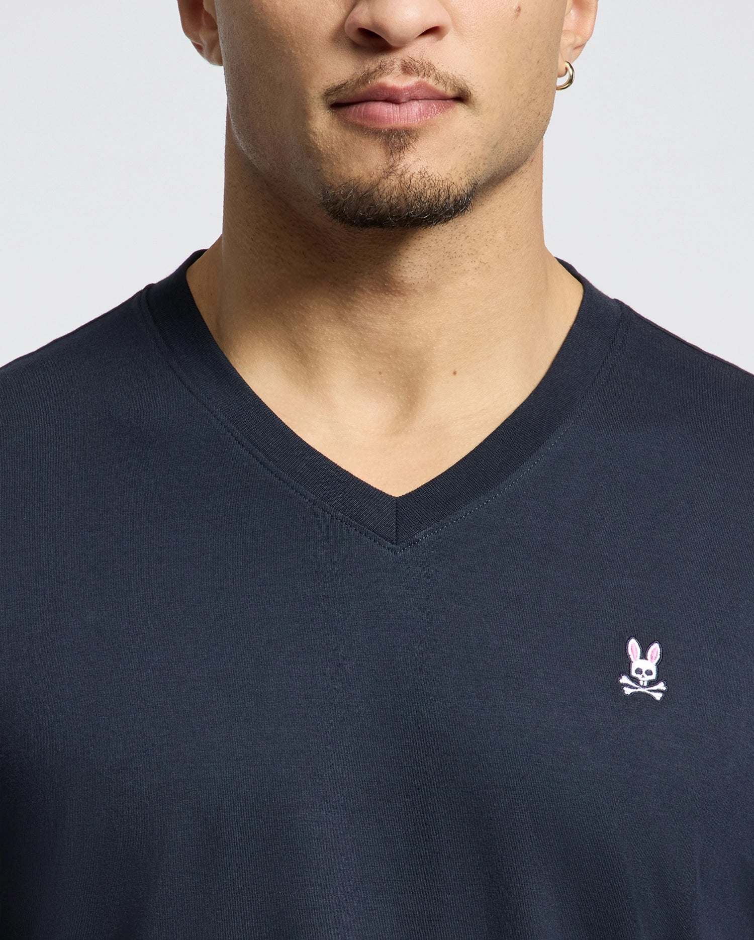 A man is wearing a MENS CLASSIC V NECK TEE - B6U100ARPC from Psycho Bunny, made of soft Pima cotton and adorned with a pink bunny and crossbones logo on the chest. He has a short beard and a small hoop earring in one ear, all set against a plain, light-colored background.