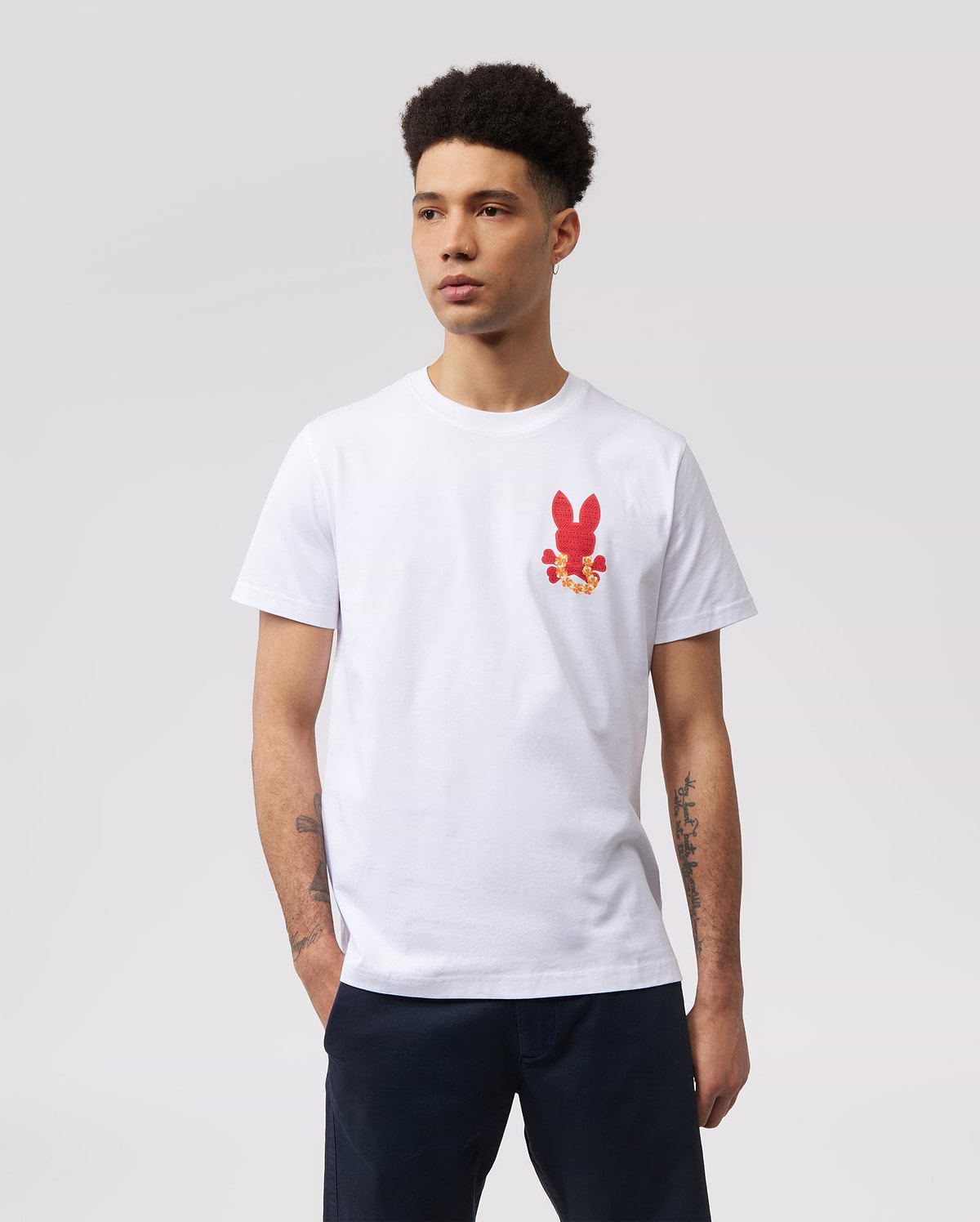 A person with short curly hair is wearing a white MENS HAWAII CITY TEE - B6U169Y1PC by Psycho Bunny made of soft Pima cotton jersey, featuring a small red bunny graphic pattern on the left side of the chest. Their hands are in their pockets, and they are standing against a plain, light gray background.