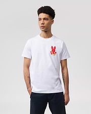 A person with short curly hair is wearing a white MENS HAWAII CITY TEE - B6U169Y1PC by Psycho Bunny made of soft Pima cotton jersey, featuring a small red bunny graphic pattern on the left side of the chest. Their hands are in their pockets, and they are standing against a plain, light gray background.