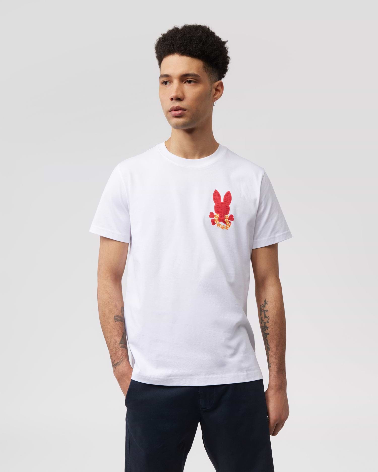 A person with short curly hair is wearing a white MENS HAWAII CITY TEE - B6U169Y1PC by Psycho Bunny made of soft Pima cotton jersey, featuring a small red bunny graphic pattern on the left side of the chest. Their hands are in their pockets, and they are standing against a plain, light gray background.