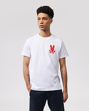 A person with short curly hair is wearing a white MENS HAWAII CITY TEE - B6U169Y1PC by Psycho Bunny made of soft Pima cotton jersey, featuring a small red bunny graphic pattern on the left side of the chest. Their hands are in their pockets, and they are standing against a plain, light gray background.