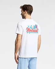 A man stands facing away from the camera in a plain setting, wearing light blue shorts and the Psycho Bunny MENS MONTREAL TEE (B6U381F200), a white T-shirt featuring colorful "Montréal" text and graphics on the back.