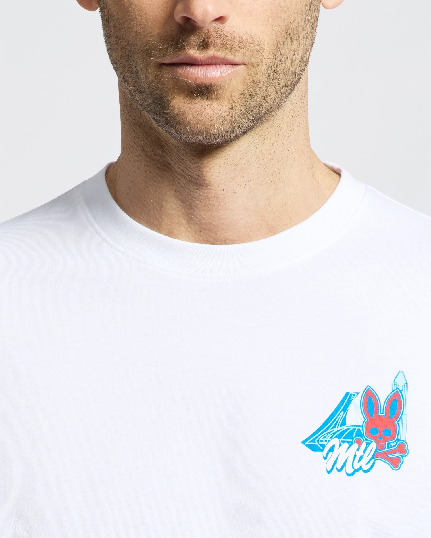 A close-up of a man in the Psycho Bunny MENS MONTREAL TEE, made from soft Pima cotton, with a small, colorful chest graphic showing the number 4, "Mil," and a red bunny face with blue ears and a blue sword.