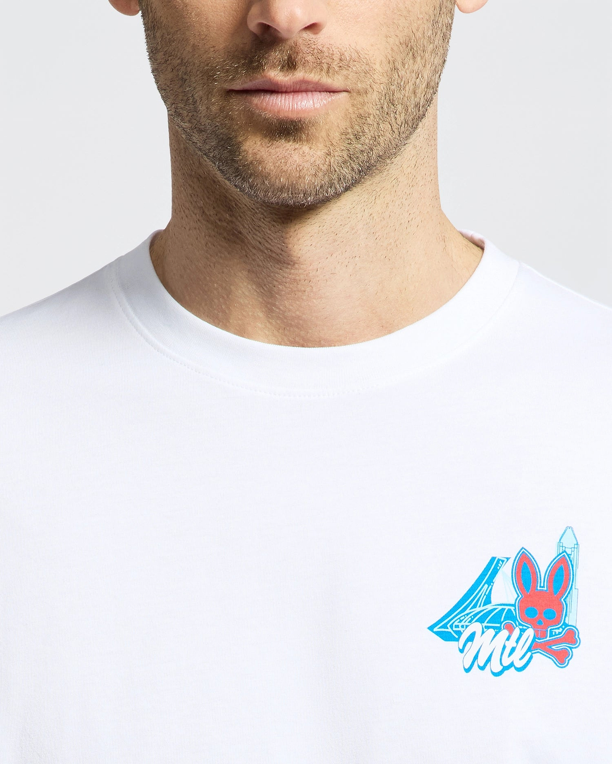 A close-up of a man in the Psycho Bunny MENS MONTREAL TEE, made from soft Pima cotton, with a small, colorful chest graphic showing the number 4, "Mil," and a red bunny face with blue ears and a blue sword.