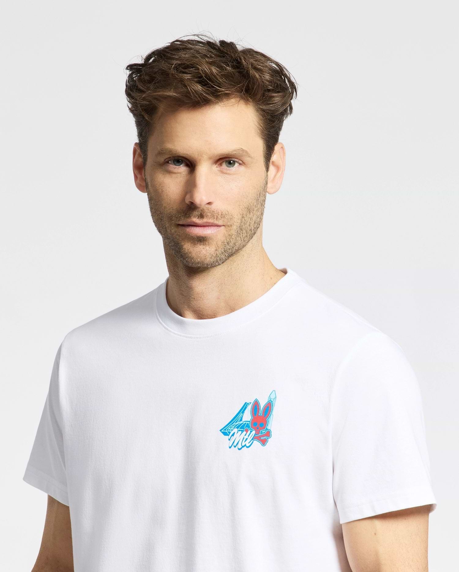 A man with short brown hair and light stubble wears the Psycho Bunny MENS MONTREAL TEE (B6U381F200), a white t-shirt featuring a small, colorful bunny graphic with a blue cape on the chest, set against a plain white background.
