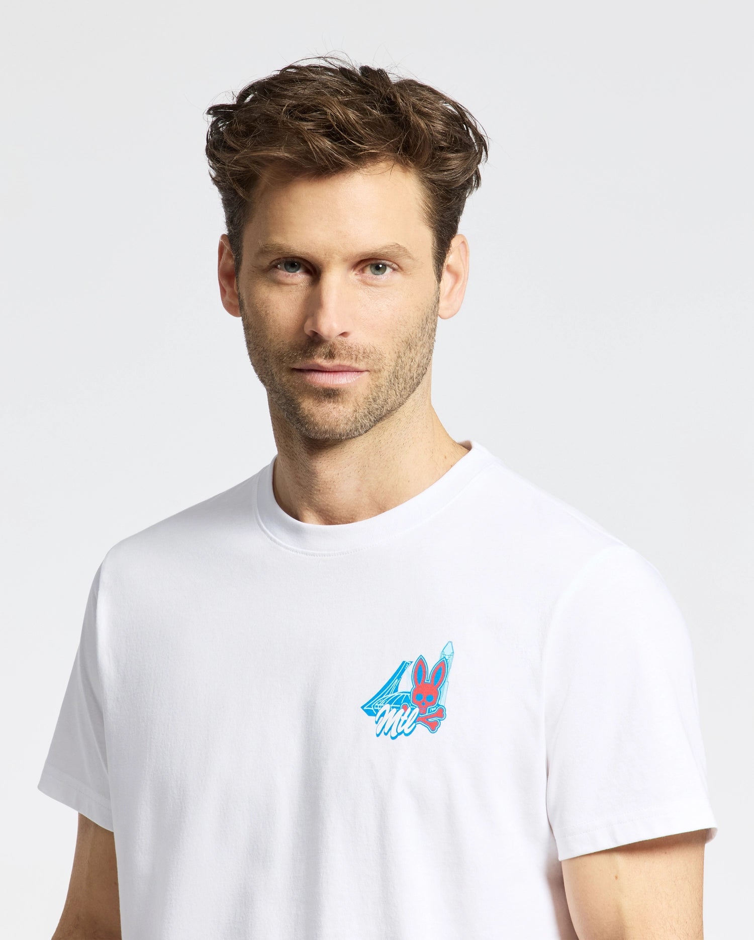 A man with short brown hair and light stubble wears the Psycho Bunny MENS MONTREAL TEE (B6U381F200), a white t-shirt featuring a small, colorful bunny graphic with a blue cape on the chest, set against a plain white background.