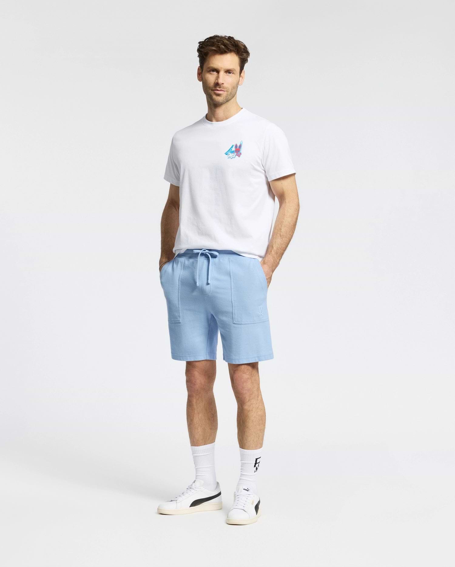 A man stands against a plain background, wearing the Psycho Bunny MENS MONTREAL TEE (B6U381F200), paired with light blue drawstring shorts, white crew socks, and white sneakers. His hands are in his pockets and he faces the camera.