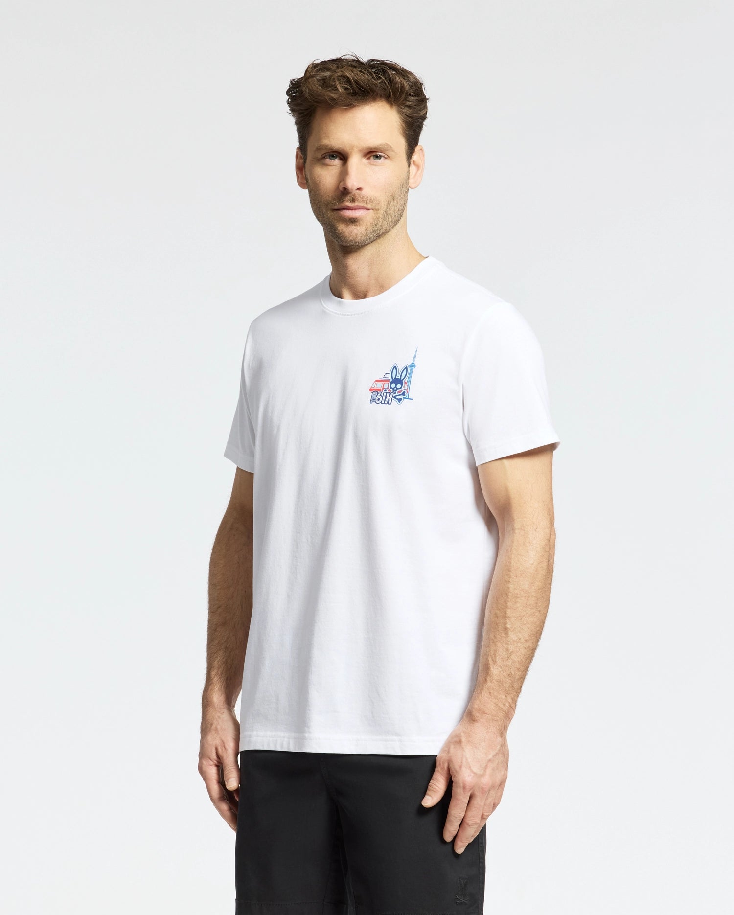 A man with short brown hair wears the Psycho Bunny MENS TORONTO TEE - B6U383F200, featuring a hometown hero graphic on the left chest, and black shorts, as he stands slightly turned against a plain white background.