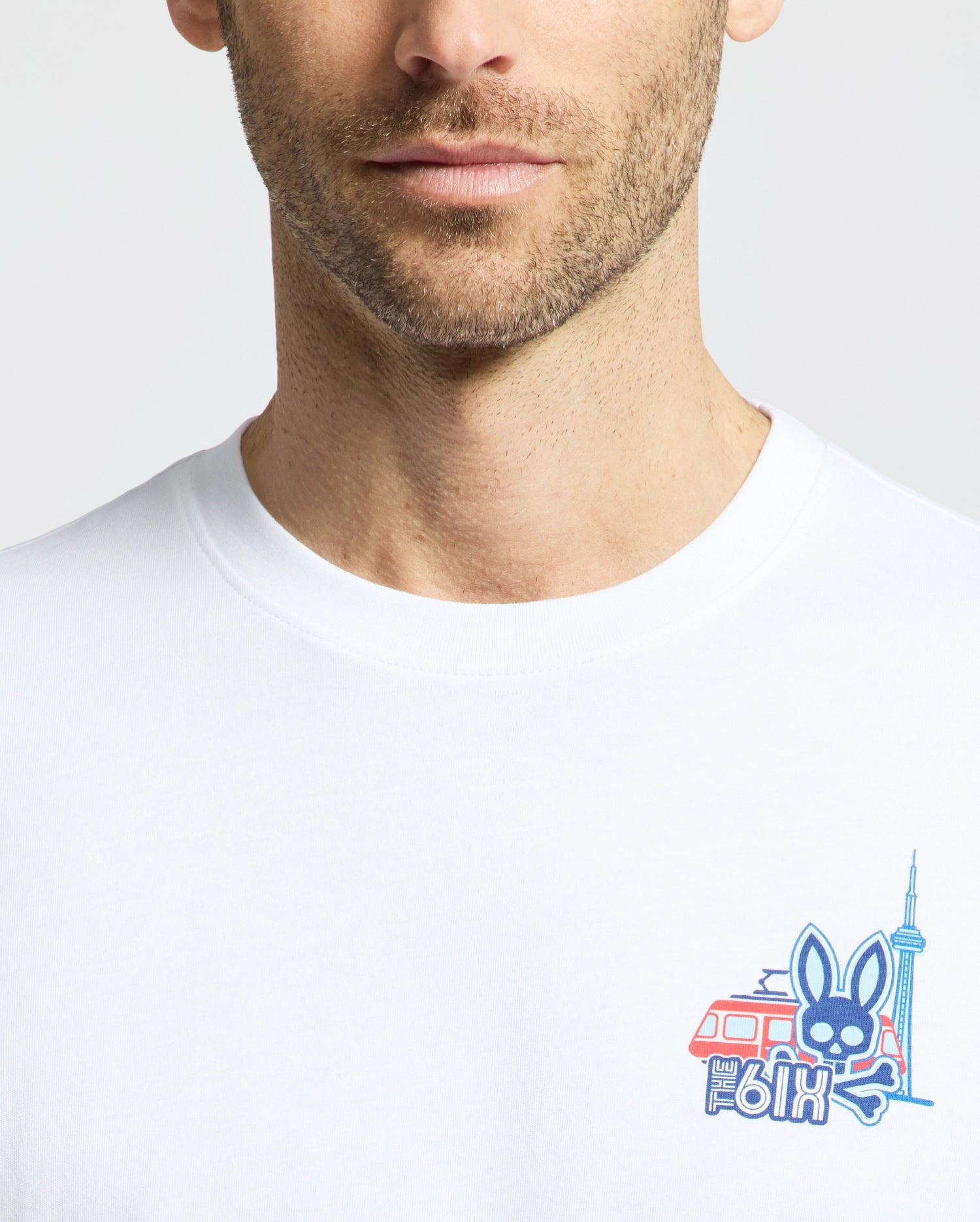 A man wears the Psycho Bunny MENS TORONTO TEE (B6U383F200), a soft Pima cotton shirt with a vibrant hometown hero graphic—bunny, streetcar, CN Tower, and "6ix"—on the chest. Only his lower face and upper torso are visible.