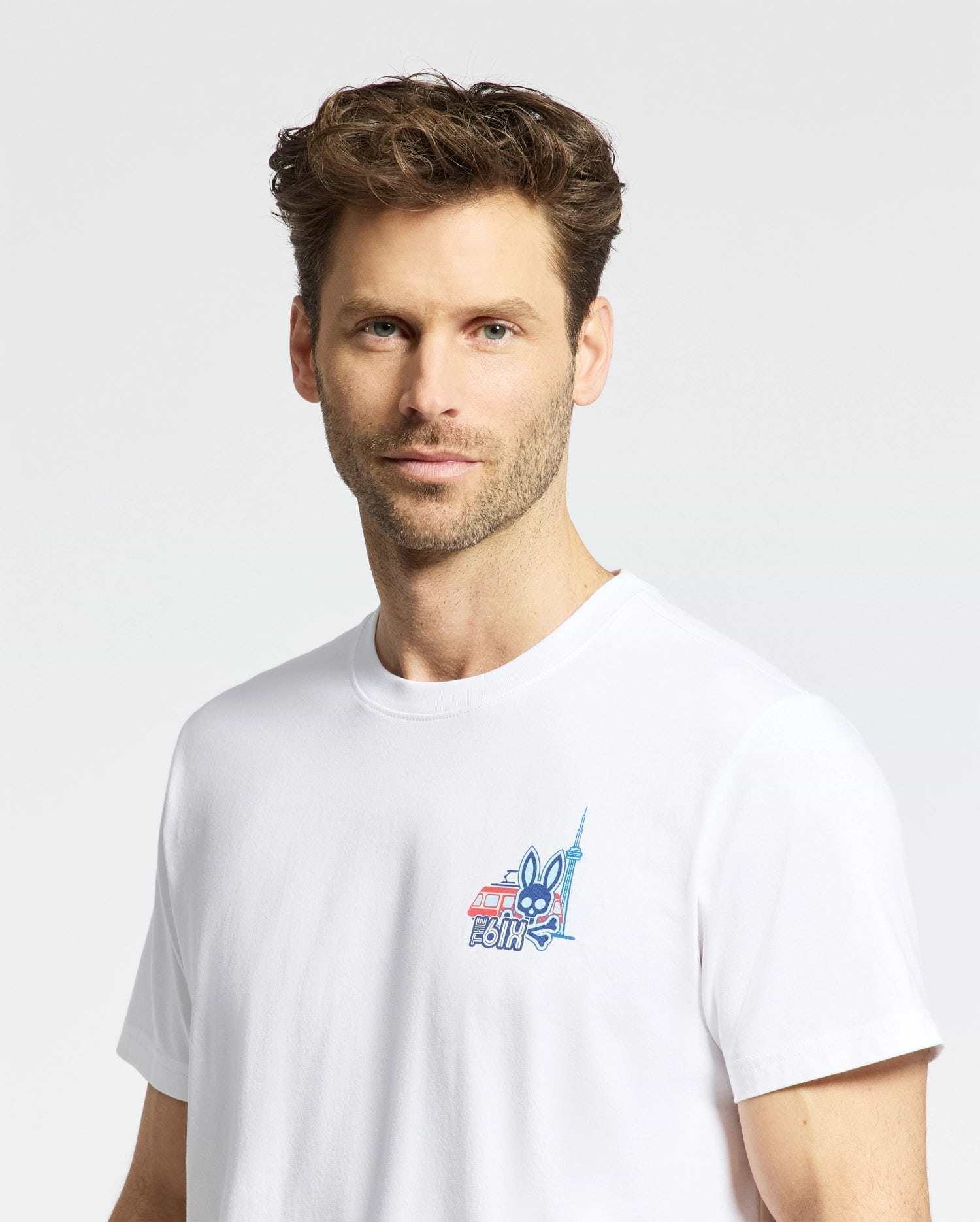 A man with short, wavy brown hair and light stubble wears the Psycho Bunny MENS TORONTO TEE - B6U383F200, a plain white Pima cotton shirt with a small colorful graphic on the chest, standing against a light background.