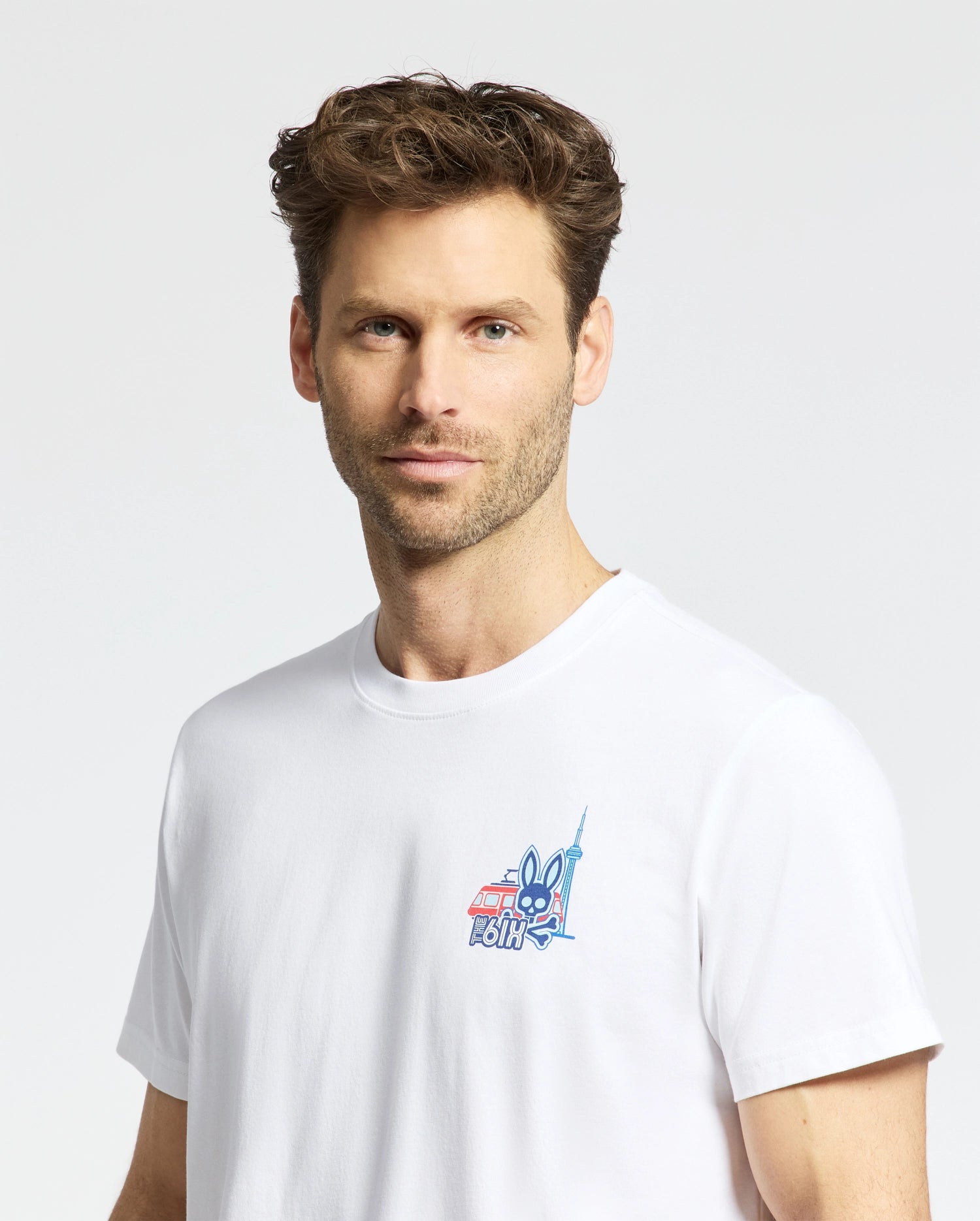 A man with short, wavy brown hair and light stubble wears the Psycho Bunny MENS TORONTO TEE - B6U383F200, a plain white Pima cotton shirt with a small colorful graphic on the chest, standing against a light background.