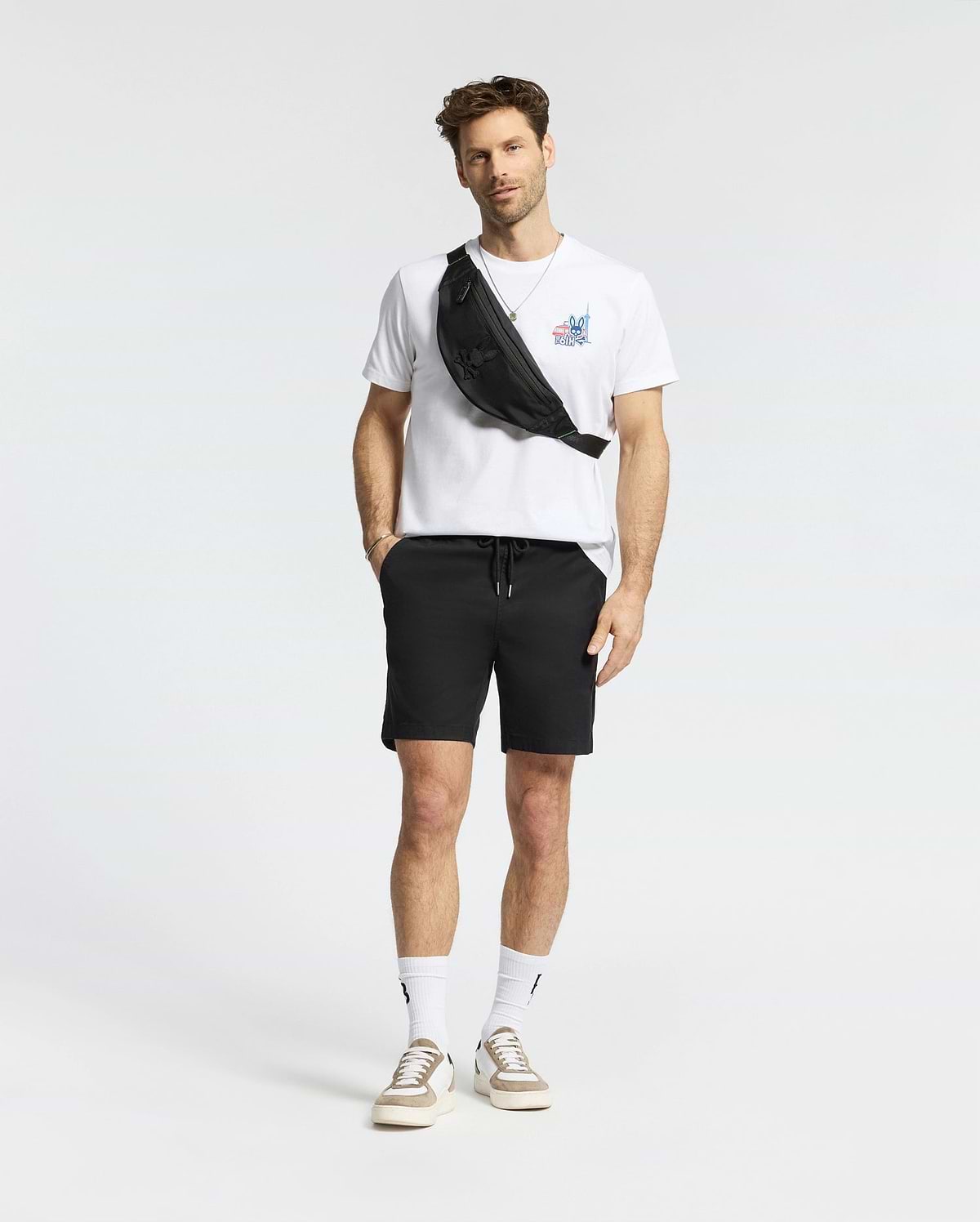 A man stands against a plain background wearing a white Psycho Bunny MENS TORONTO TEE (B6U383F200), black shorts, white socks, beige sneakers, and a black crossbody bag.