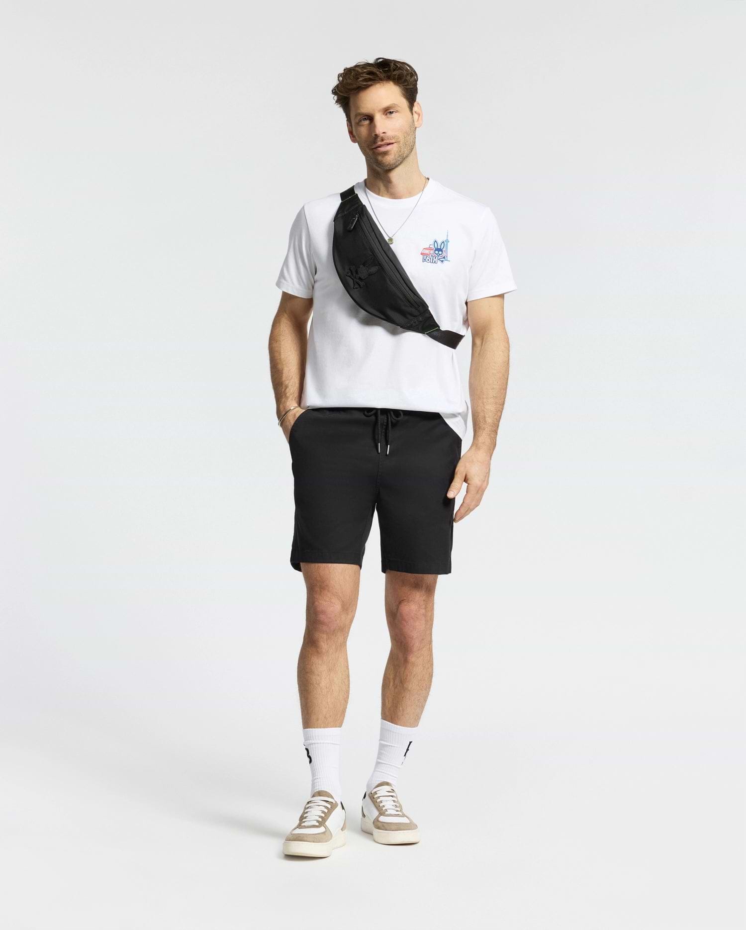 A man stands against a plain background wearing a white Psycho Bunny MENS TORONTO TEE (B6U383F200), black shorts, white socks, beige sneakers, and a black crossbody bag.