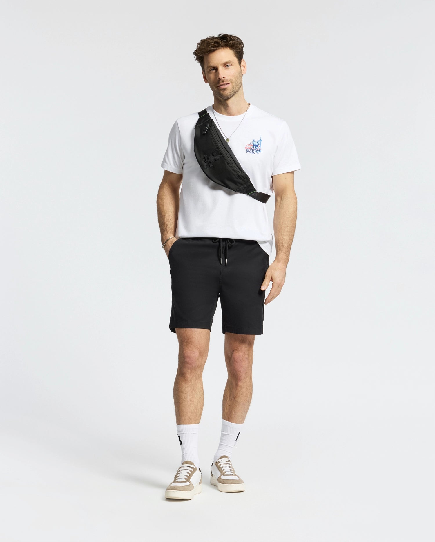 A man stands against a plain background wearing a white Psycho Bunny MENS TORONTO TEE (B6U383F200), black shorts, white socks, beige sneakers, and a black crossbody bag.