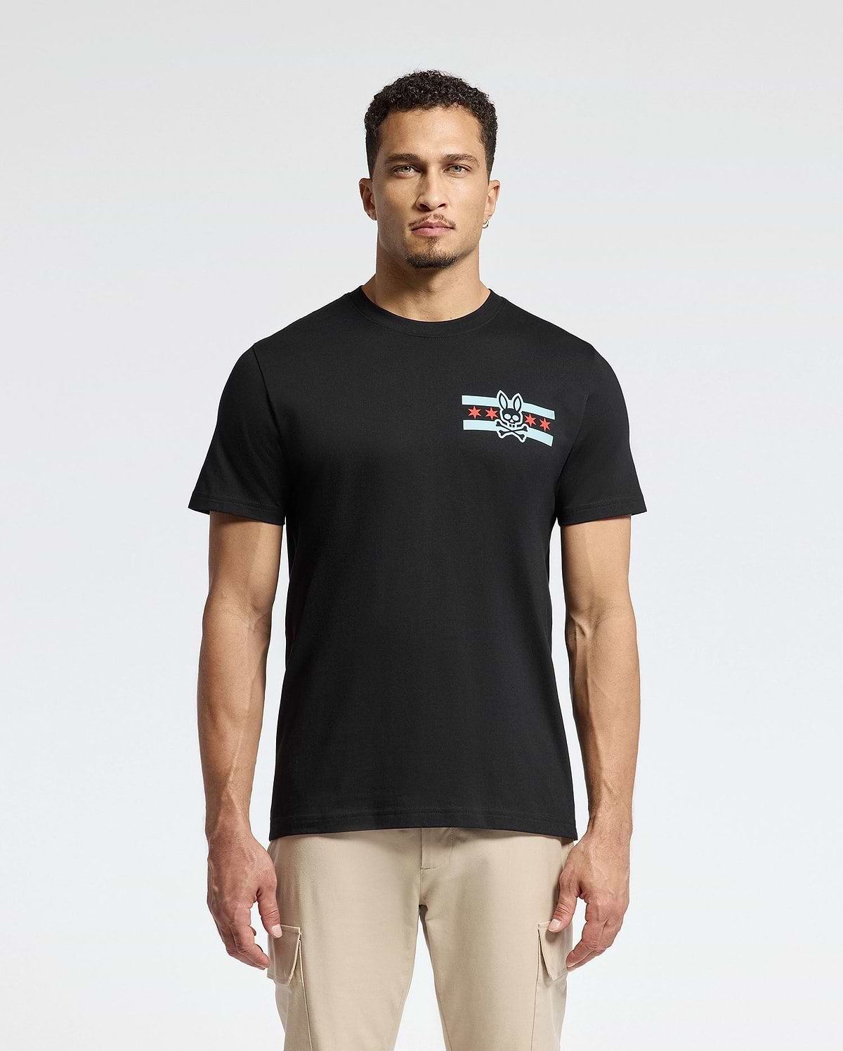 A man wearing the Psycho Bunny MENS CHICAGO TEE - B6U551W1PC, crafted from soft Pima cotton and featuring a small graphic design with stripes and a rabbit emblem on the left chest, is complemented by light beige pants as he stands confidently against a plain white background.