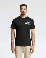 A man wearing the Psycho Bunny MENS CHICAGO TEE - B6U551W1PC, crafted from soft Pima cotton and featuring a small graphic design with stripes and a rabbit emblem on the left chest, is complemented by light beige pants as he stands confidently against a plain white background.