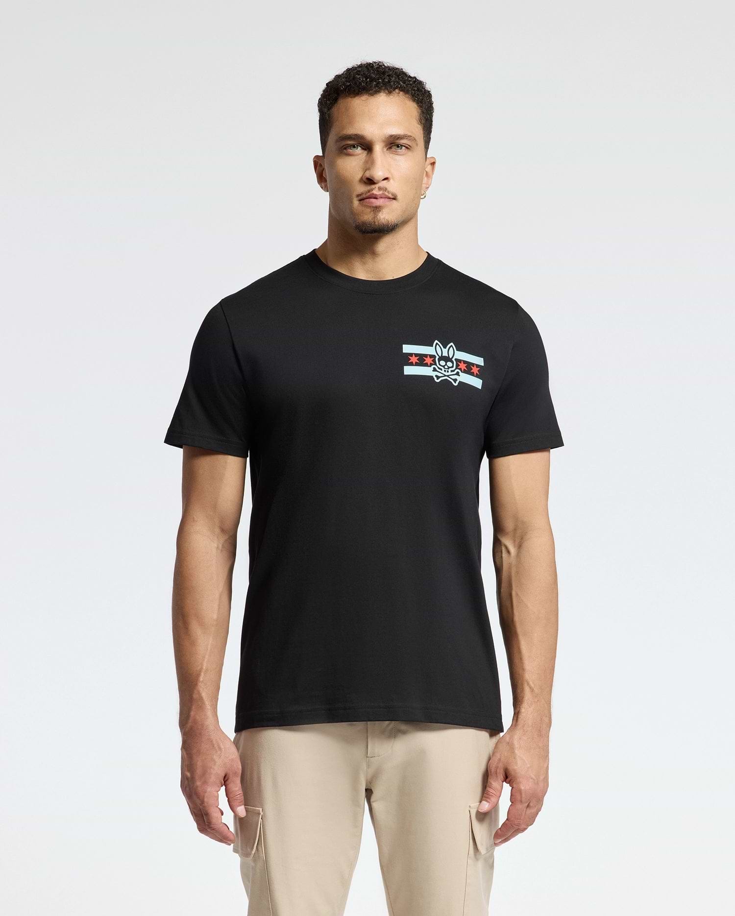 A man wearing the Psycho Bunny MENS CHICAGO TEE - B6U551W1PC, crafted from soft Pima cotton and featuring a small graphic design with stripes and a rabbit emblem on the left chest, is complemented by light beige pants as he stands confidently against a plain white background.
