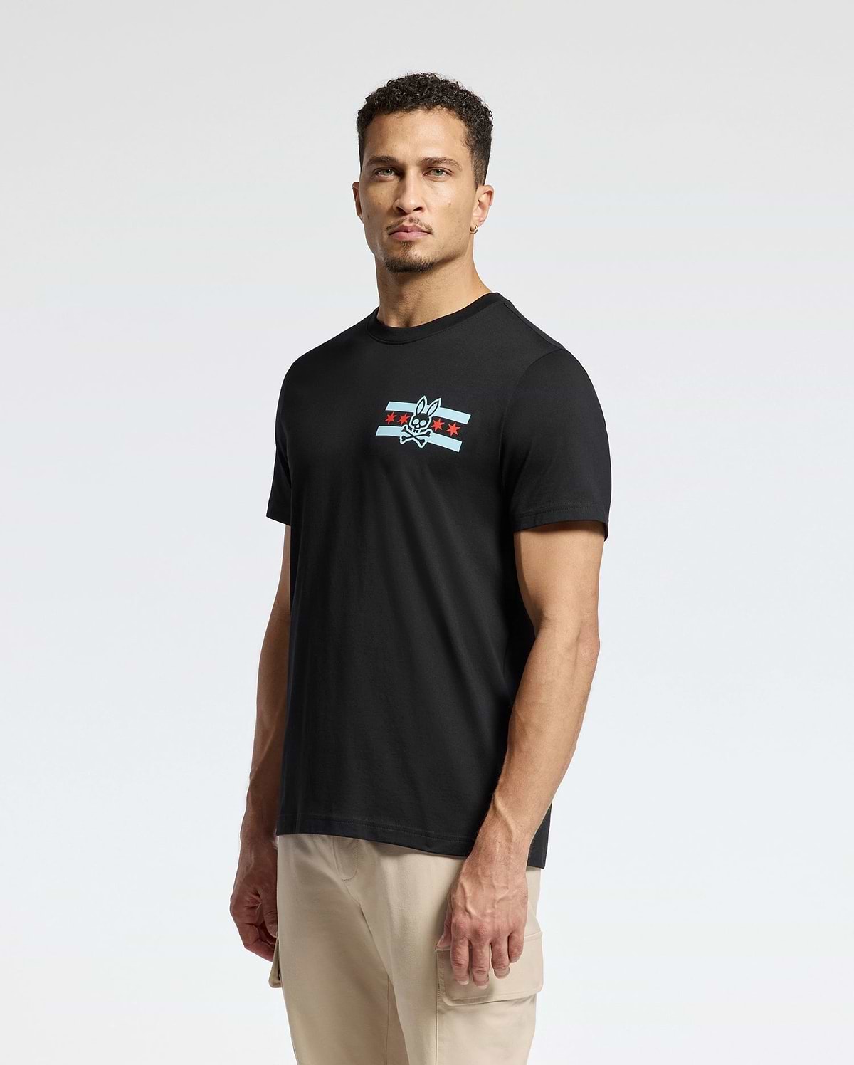 A man is standing against a plain background, wearing the Psycho Bunny MENS CHICAGO TEE - B6U551W1PC in black. This Pima cotton shirt features a small logo on the chest with a cartoonish bird and star design. He has short hair and looks directly at the camera, pairing it with light-colored pants.