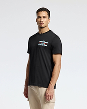 A man is standing against a plain background, wearing the Psycho Bunny MENS CHICAGO TEE - B6U551W1PC in black. This Pima cotton shirt features a small logo on the chest with a cartoonish bird and star design. He has short hair and looks directly at the camera, pairing it with light-colored pants.