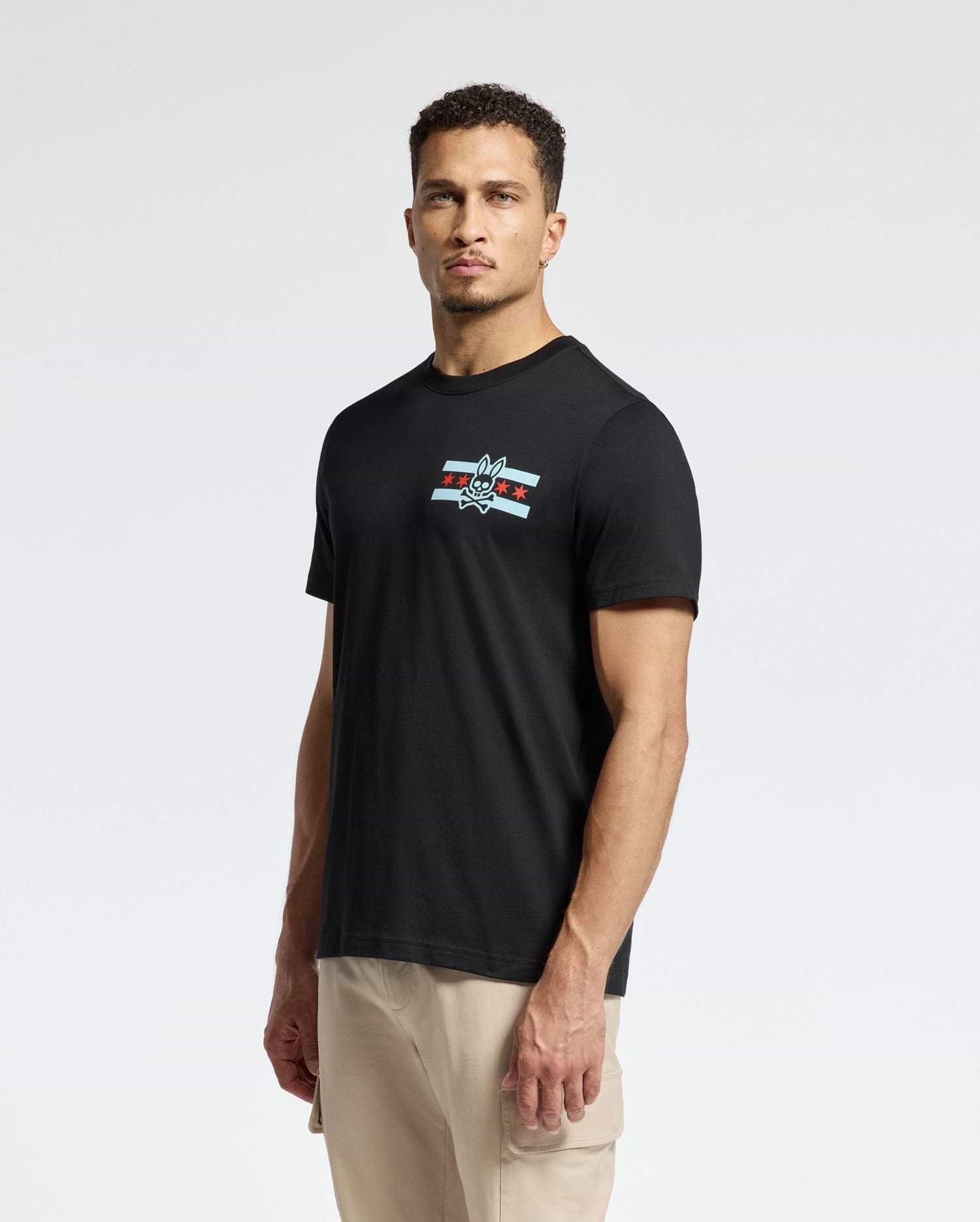 A man is standing against a plain background, wearing the Psycho Bunny MENS CHICAGO TEE - B6U551W1PC in black. This Pima cotton shirt features a small logo on the chest with a cartoonish bird and star design. He has short hair and looks directly at the camera, pairing it with light-colored pants.