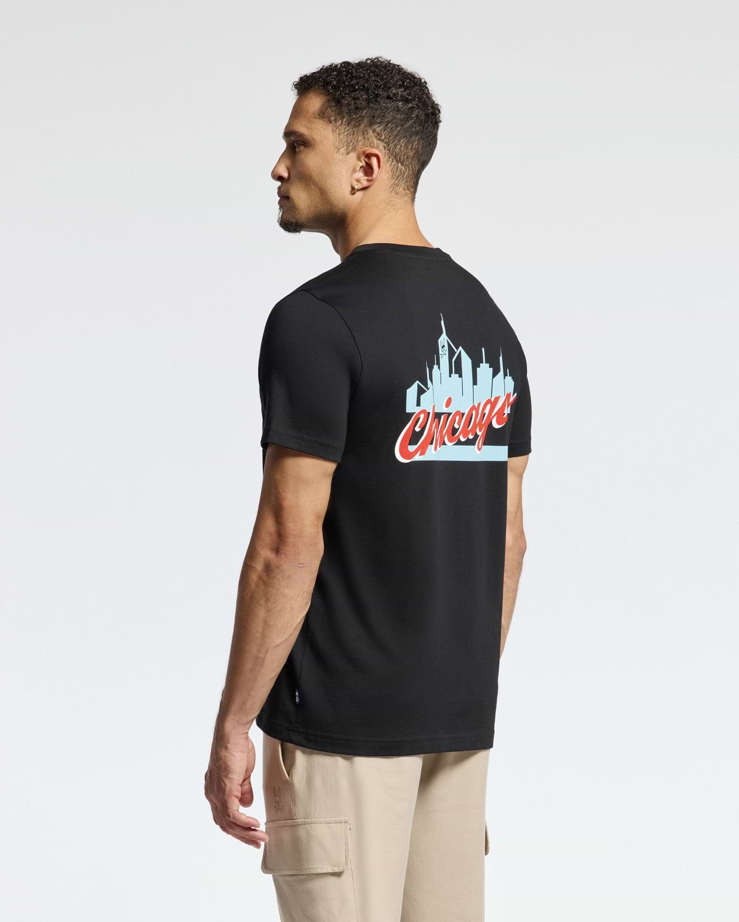 A man wearing a Psycho Bunny MENS CHICAGO TEE - B6U551W1PC crafted from soft Pima cotton is seen standing sideways against a plain background. The black tee prominently showcases a light blue skyline graphic with "Chicago" written in red cursive on the back, paired with beige cargo pants to complete his look.