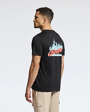 A man wearing a Psycho Bunny MENS CHICAGO TEE - B6U551W1PC crafted from soft Pima cotton is seen standing sideways against a plain background. The black tee prominently showcases a light blue skyline graphic with "Chicago" written in red cursive on the back, paired with beige cargo pants to complete his look.