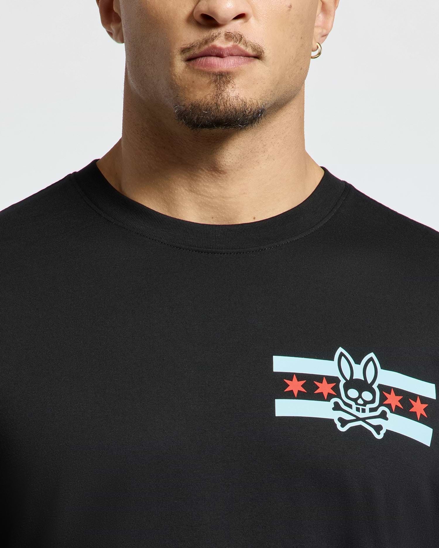 A person wearing a stylish Men's Chicago Tee by Psycho Bunny in black, featuring a graphic design of a bunny skull and crossbones surrounded by blue stripes and red stars. The image highlights the upper portion of this shirt.