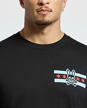 A person wearing a stylish Men's Chicago Tee by Psycho Bunny in black, featuring a graphic design of a bunny skull and crossbones surrounded by blue stripes and red stars. The image highlights the upper portion of this shirt.