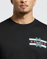 A person wearing a stylish Men's Chicago Tee by Psycho Bunny in black, featuring a graphic design of a bunny skull and crossbones surrounded by blue stripes and red stars. The image highlights the upper portion of this shirt.