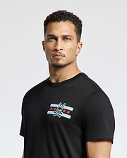 A man with short curly hair is wearing the Psycho Bunny MENS CHICAGO TEE - B6U551W1PC, crafted from soft Pima cotton, showcasing a rabbit skull and crossbones graphic over a stylized flag with red stars and blue stripes. The background is plain white.